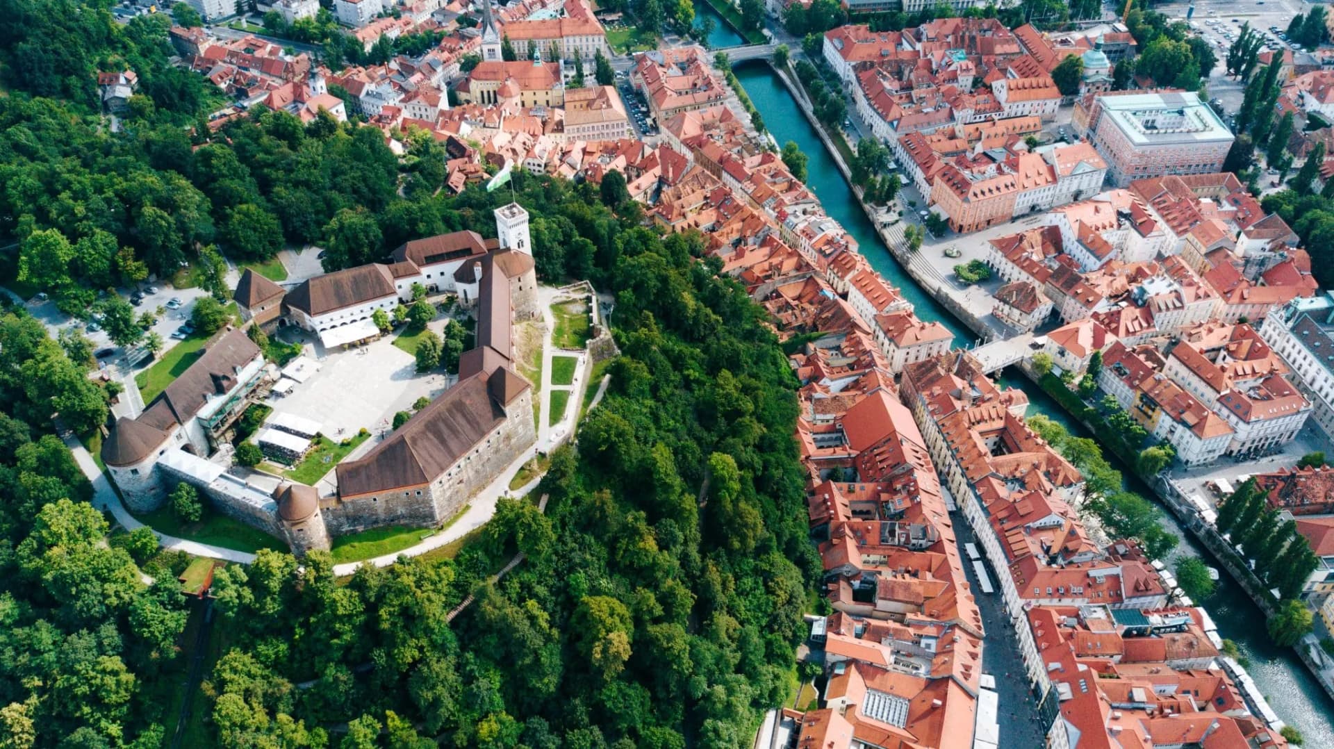 Aerial view of Ljubljana Castle overlooking dense green forest and red-roofed city buildings beside a turquoise river.