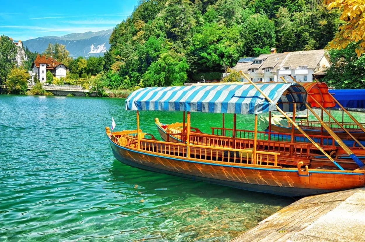 Traditional wooden Pletna boat docked on emerald water of Lake Bled with forested hills.