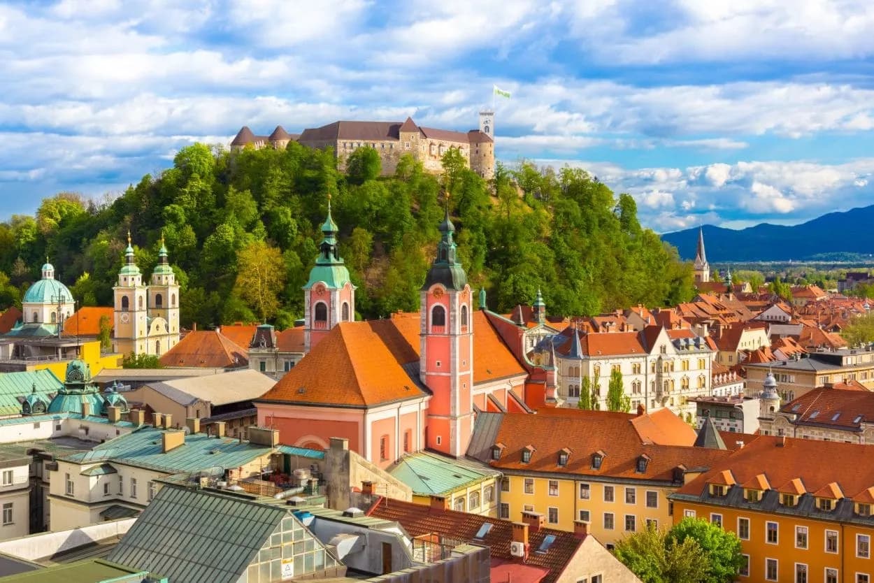 Ljubljana Castle overlooking colorful rooftops, churches, and green hills under a cloudy sky.