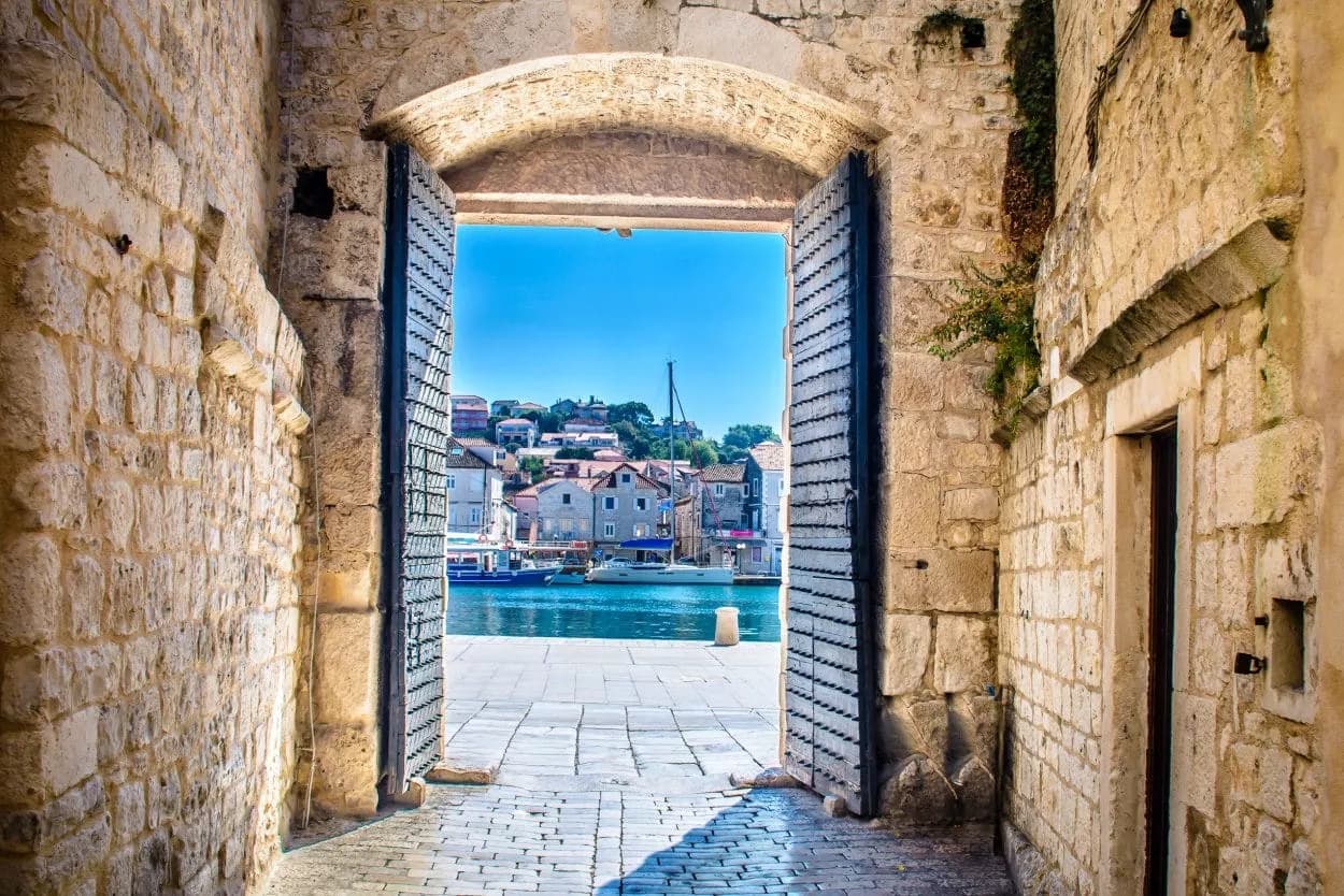 Stone archway opening to Trogir harbor with boats and colorful hillside houses.
