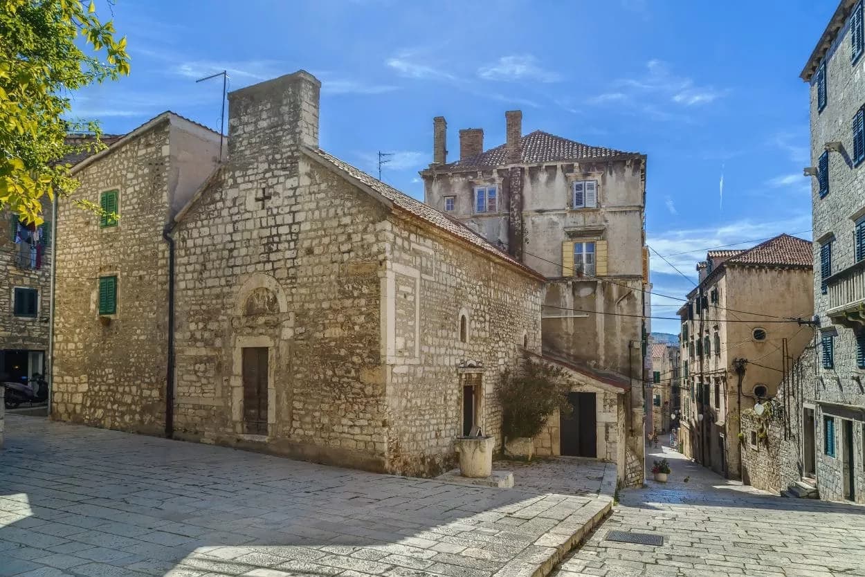 Stone church and historic buildings on a cobblestone street in Šibenik under a bright blue sky.