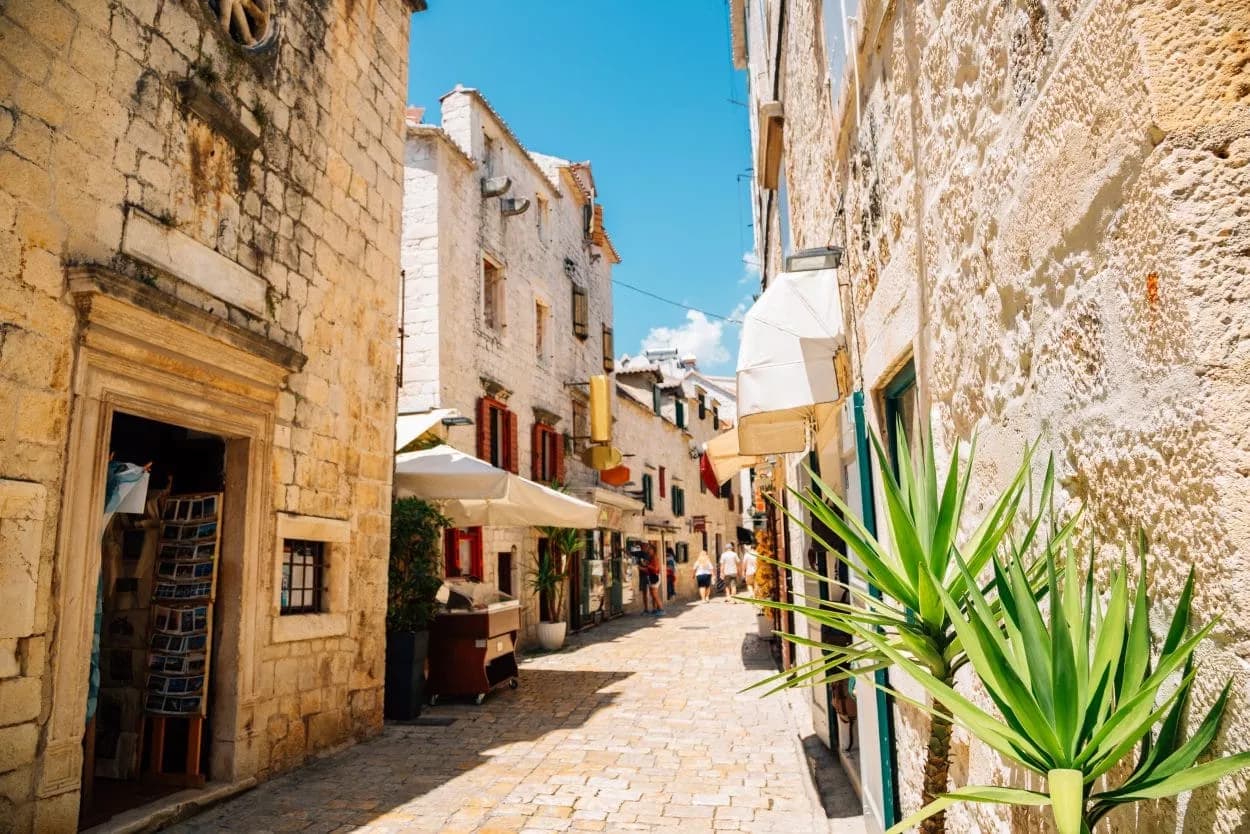 Cobblestone street in Trogir with stone buildings, awnings, and bright green plants under a blue sky.