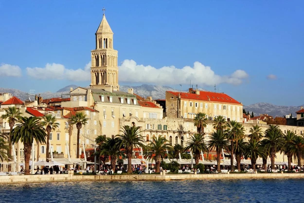 Historic waterfront with palm trees, bell tower, and snow-capped mountains in Split, Croatia.