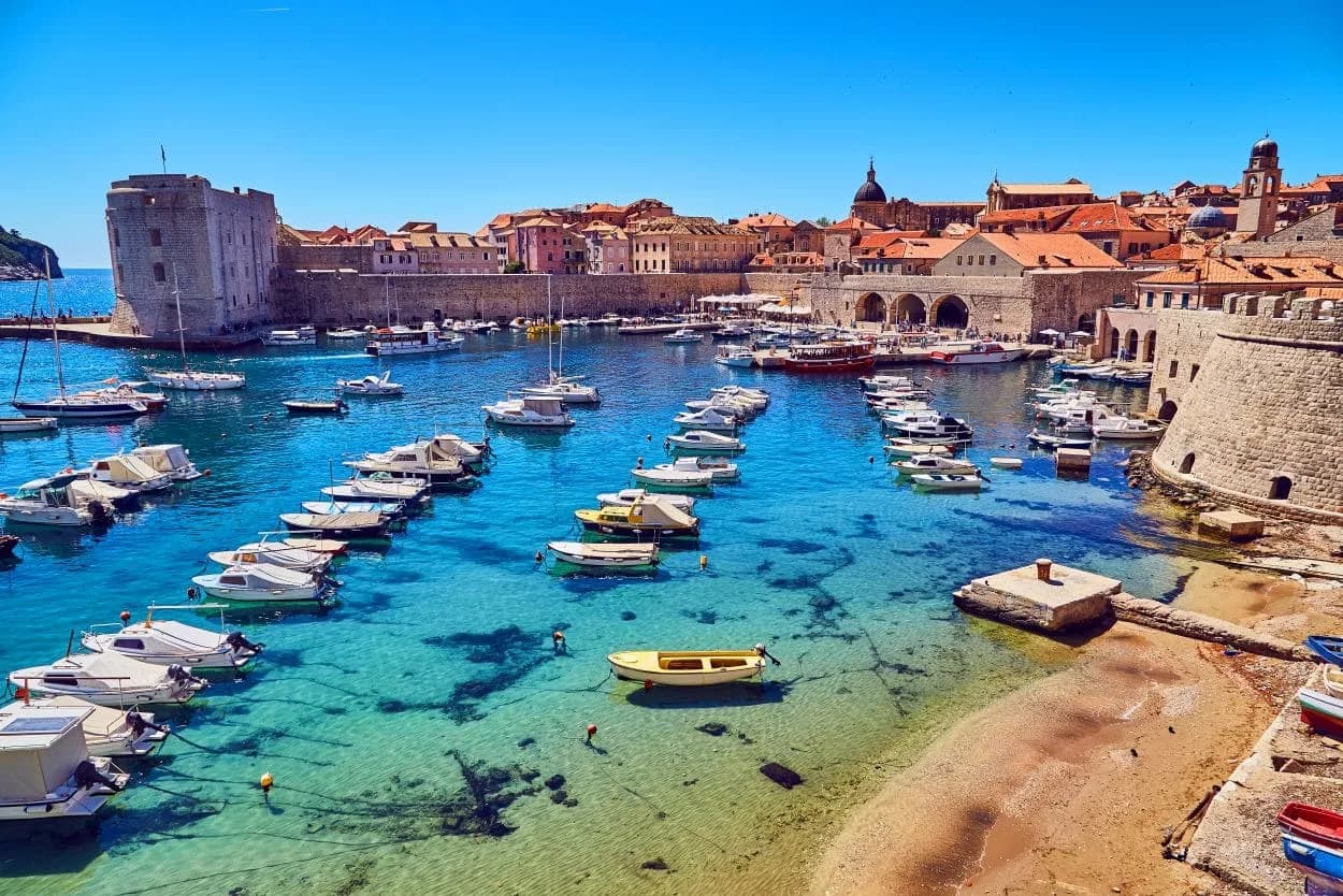 Numerous boats in clear blue water of Dubrovnik marina next to historic stone walls.