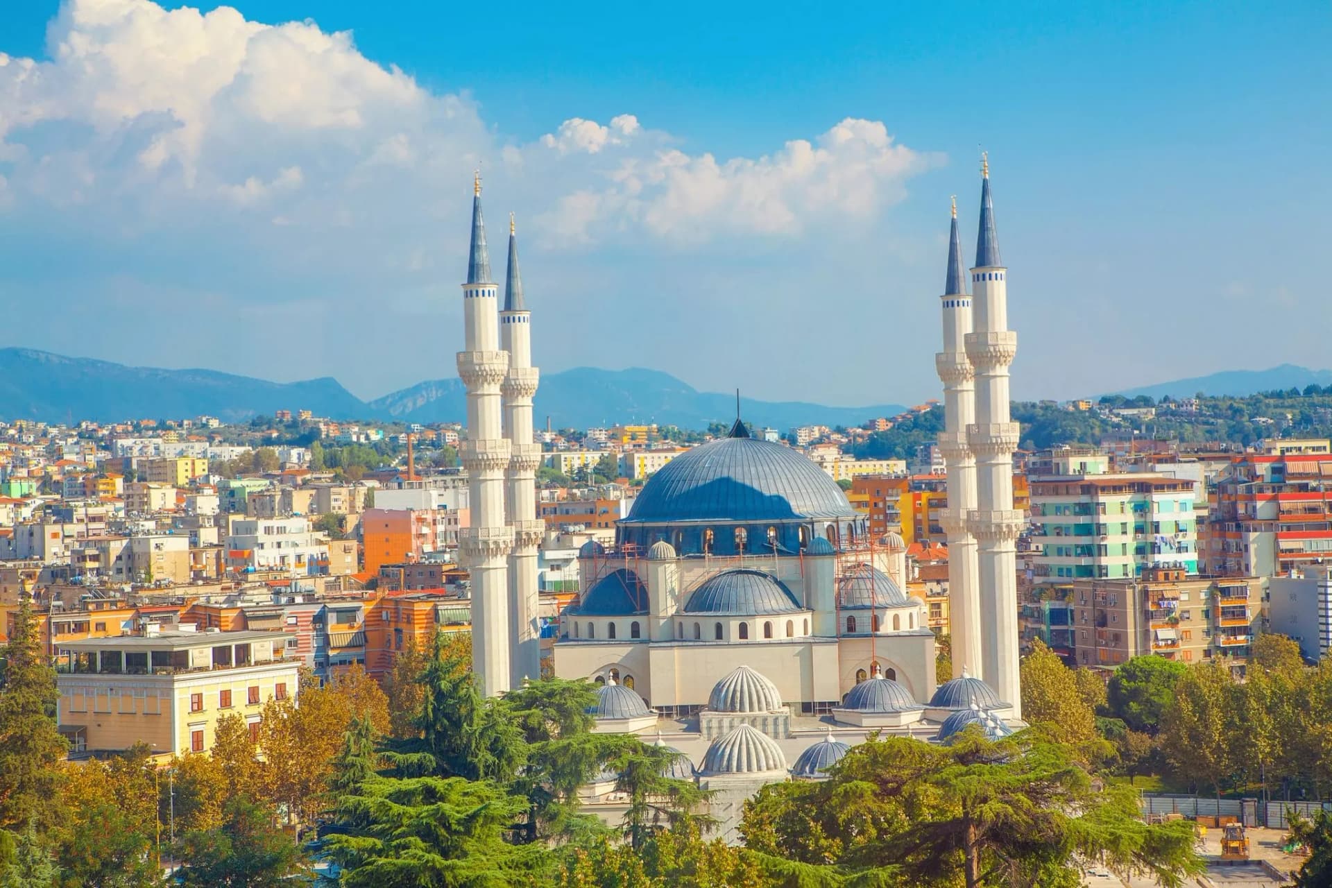 Large mosque with blue dome and twin minarets overlooking Tirana cityscape and mountains.