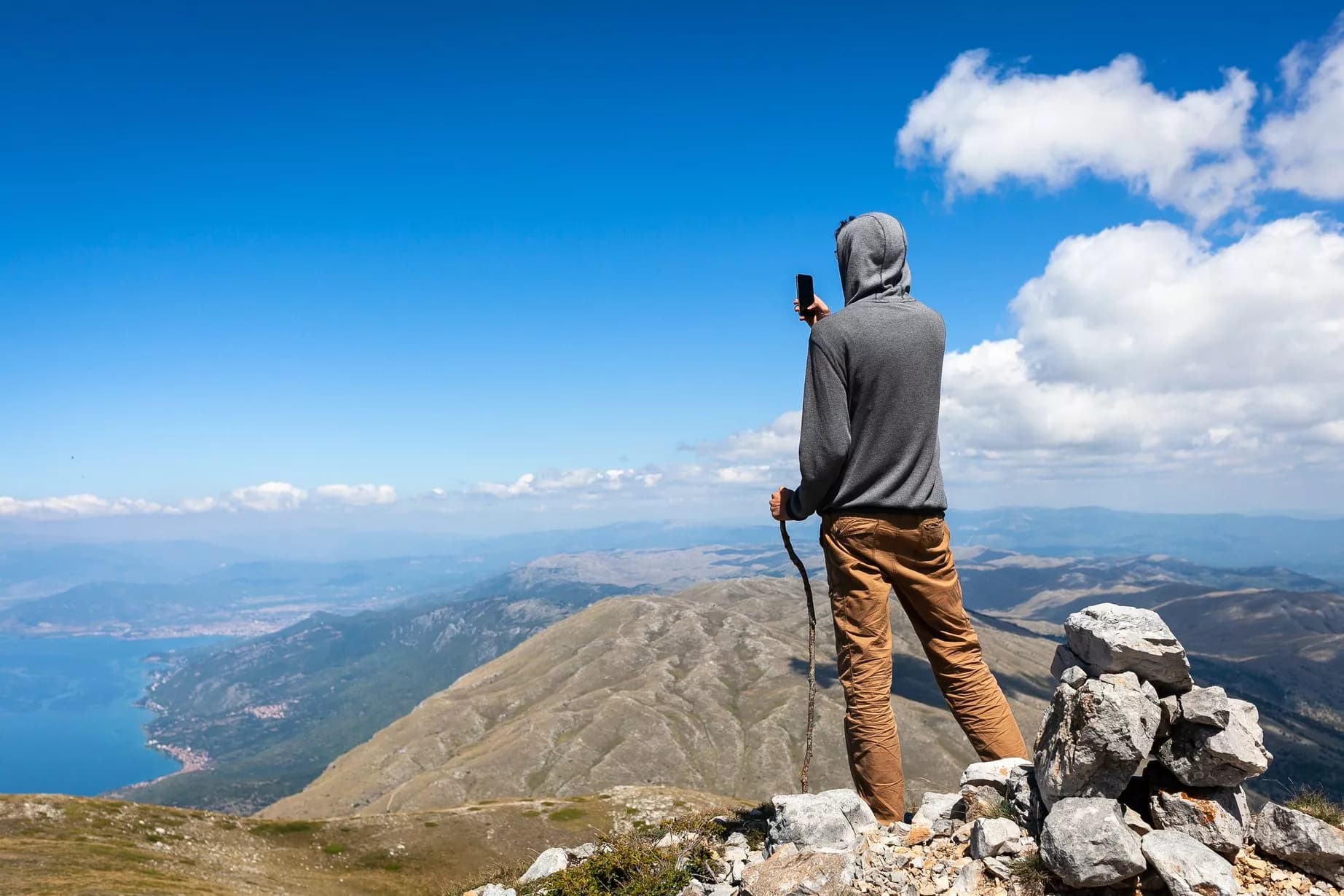 Hiker taking photo with phone atop rocky mountain overlooking vast valley and blue lake.