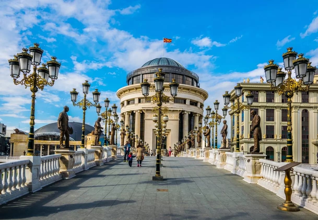 Statues and ornate lampposts line the Skopje Art Bridge under a bright blue sky.