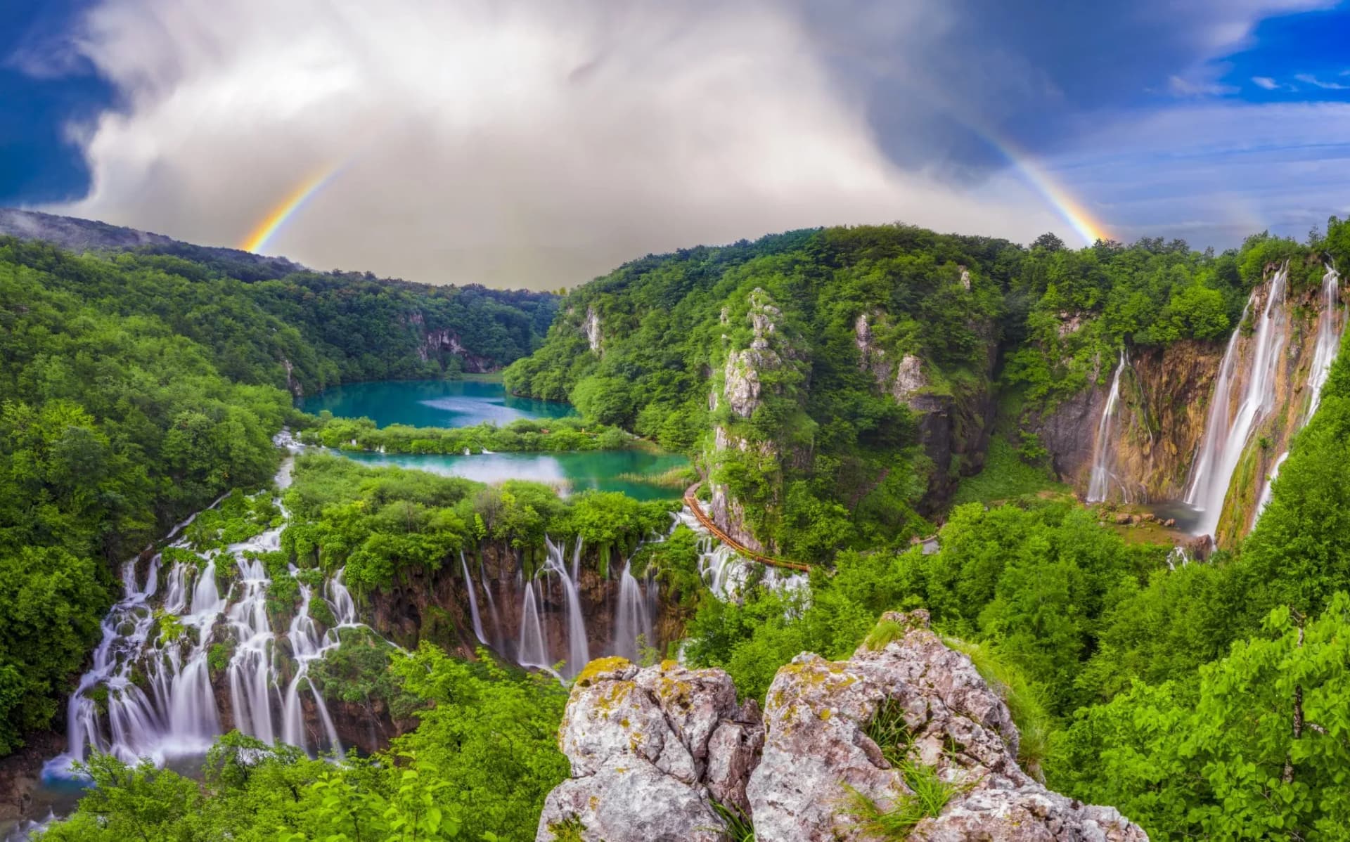 Waterfalls cascade into turquoise lakes surrounded by green forest under a rainbow at Plitvice Lakes.