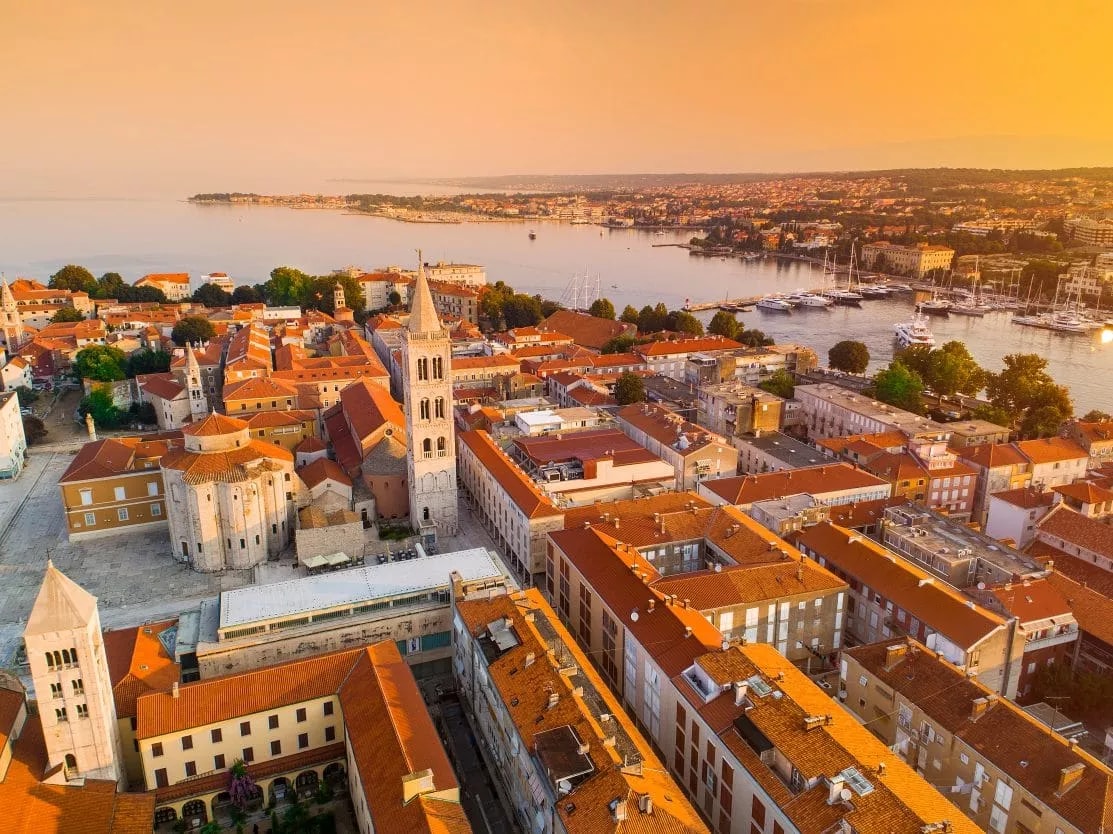 Aerial view of Zadar rooftops, bell tower, and harbor at sunset over the Adriatic Sea.
