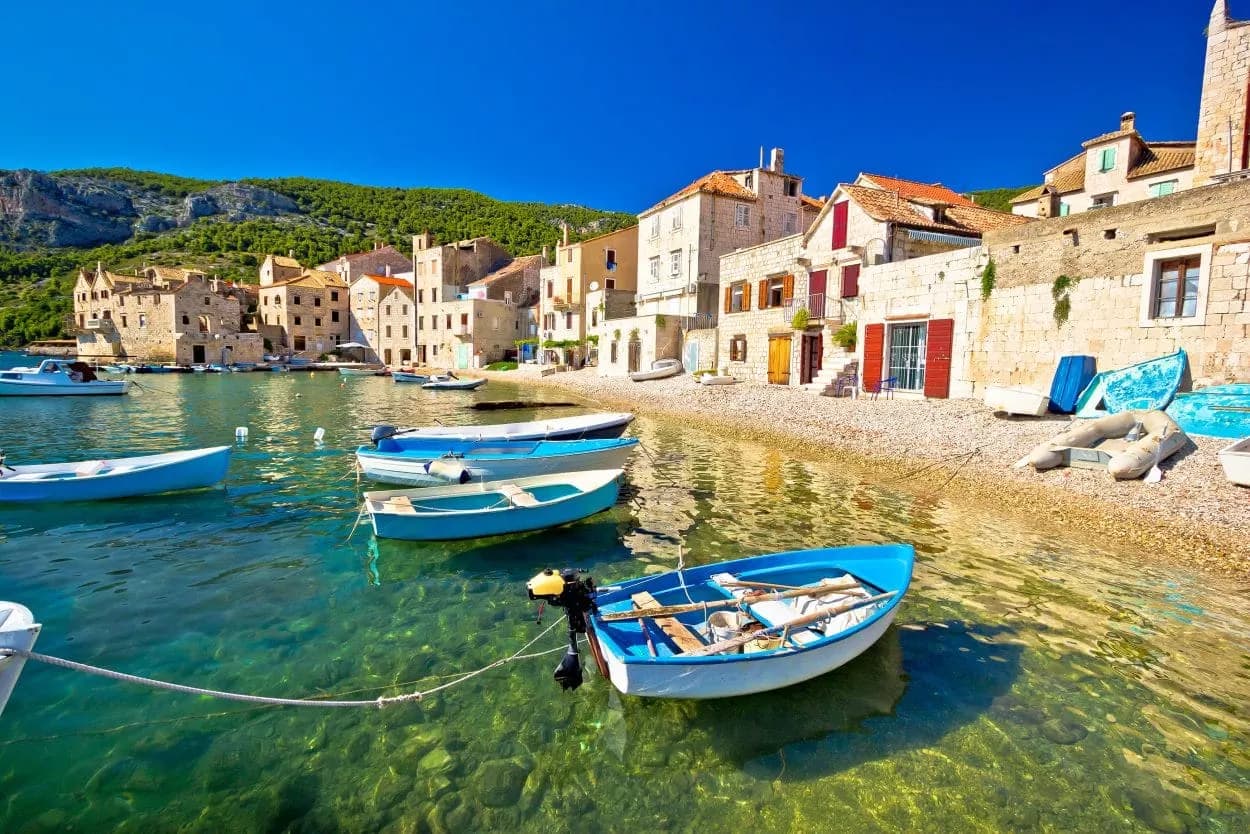 Small blue boats moored in clear turquoise water near a stone village on Vis island.