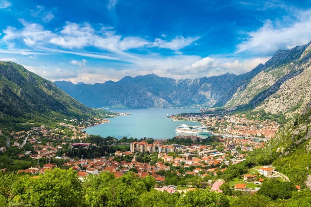 Bay of Kotor panorama with cruise ship, coastal town, and steep green mountains under blue sky.