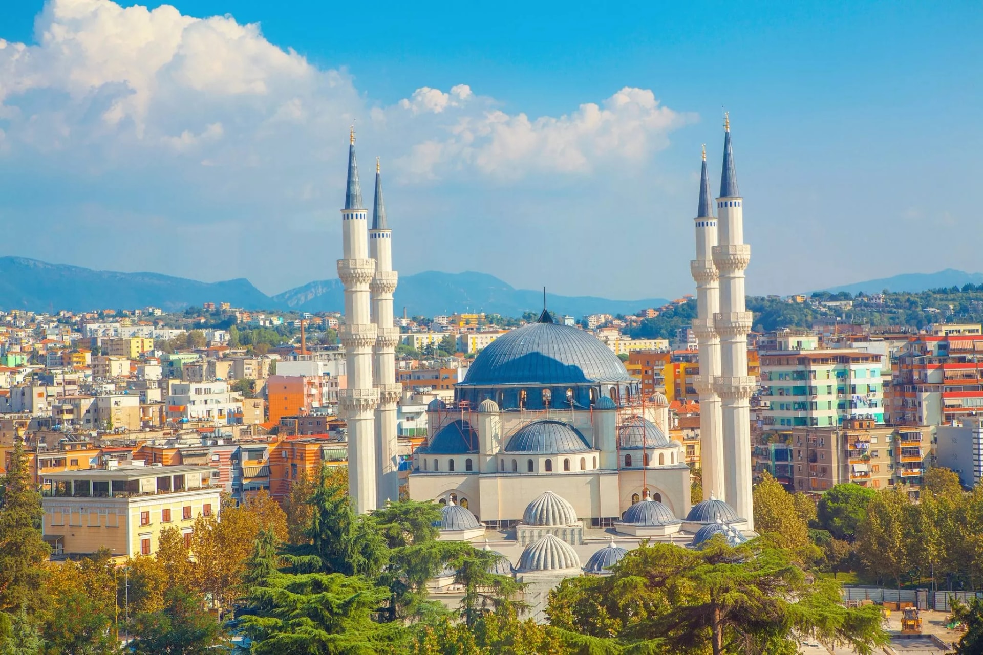 Large mosque with blue dome and minarets overlooking colorful city buildings and distant mountains in Tirana.