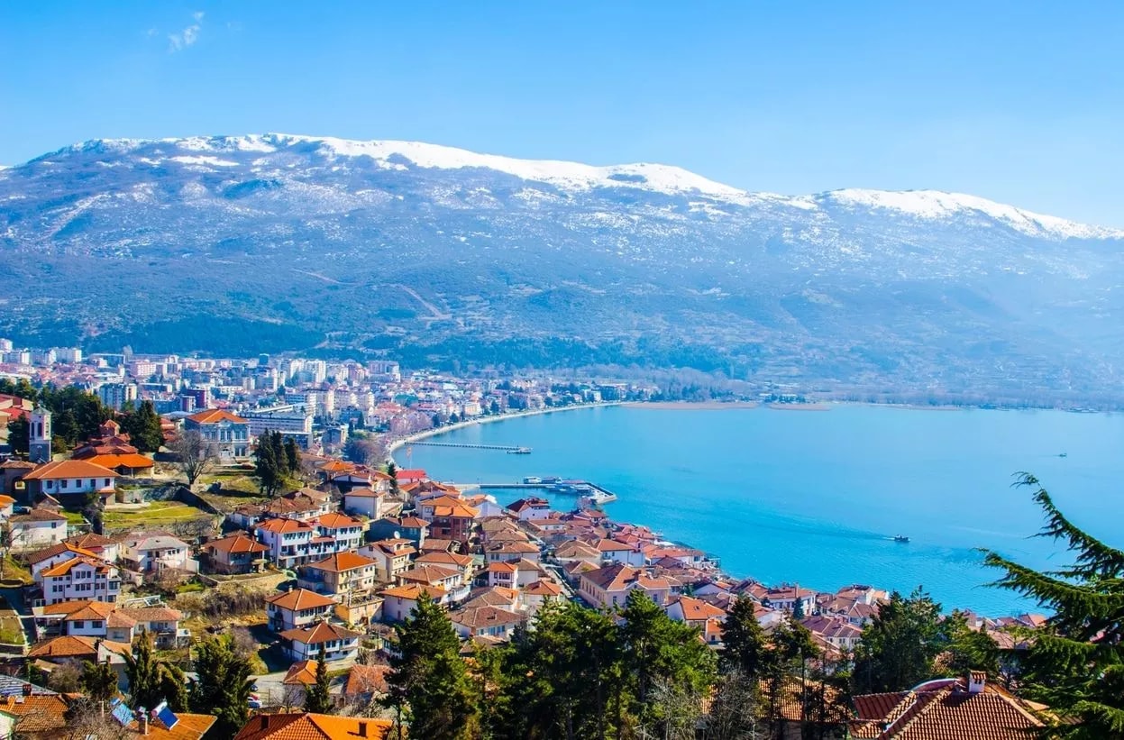 Ohrid town with terracotta roofs along bright blue lake beneath snow-capped mountains.