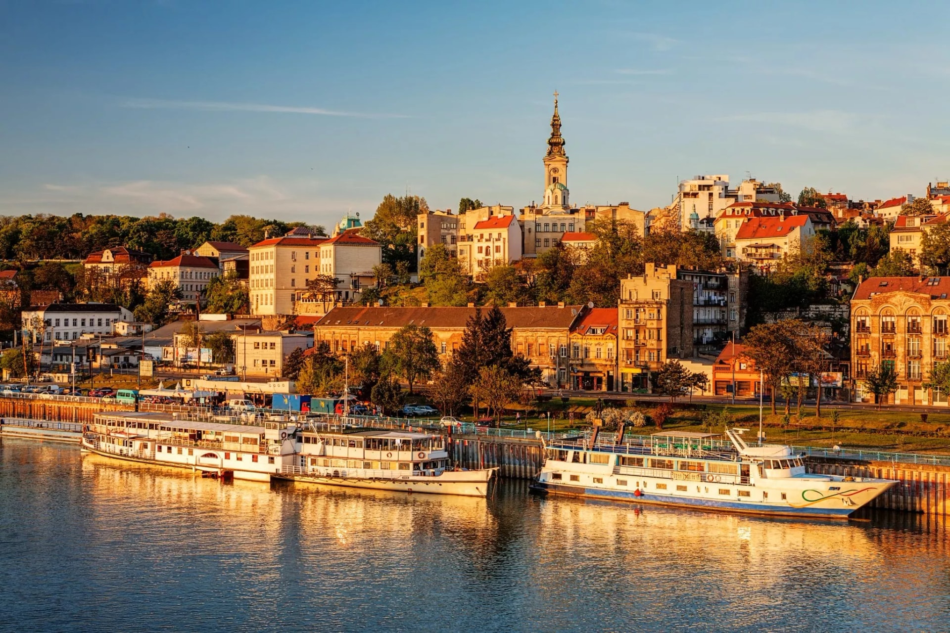 Boats docked on the Danube River with Belgrade cityscape and church tower at sunset.