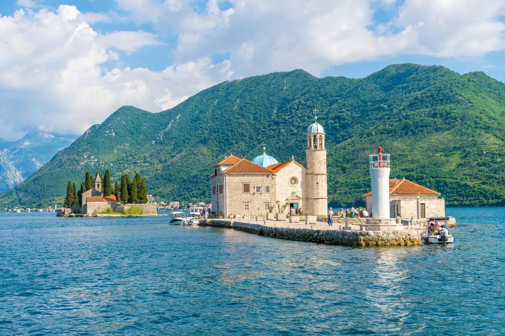Tourists sail to Our Lady of the Rocks island in Montenegro's Boka Bay with green mountains behind.