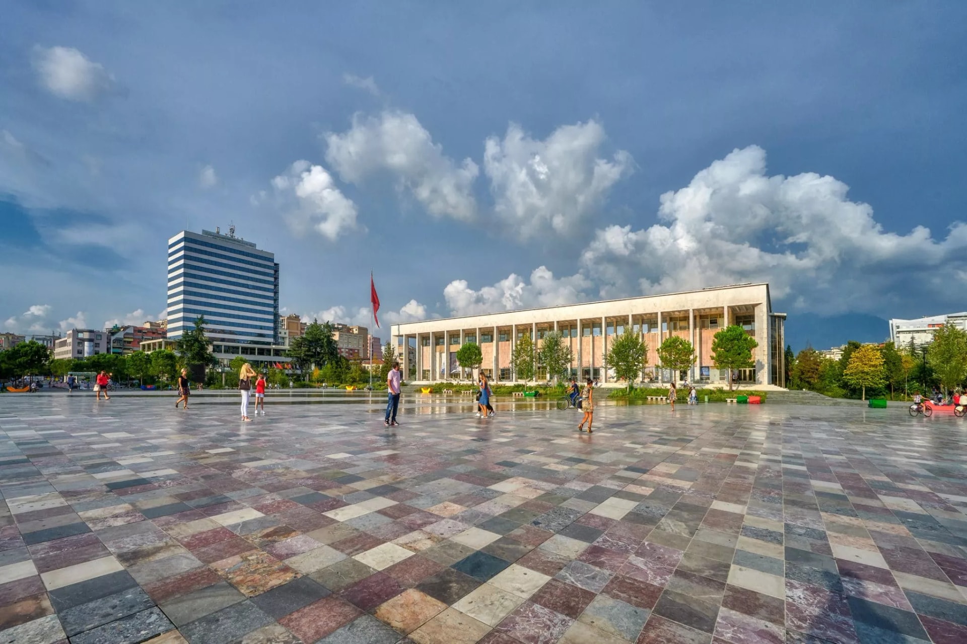 People walking across the large, multi-colored tiled Skanderbeg Square in Tirana with modern buildings under a cloudy sky.