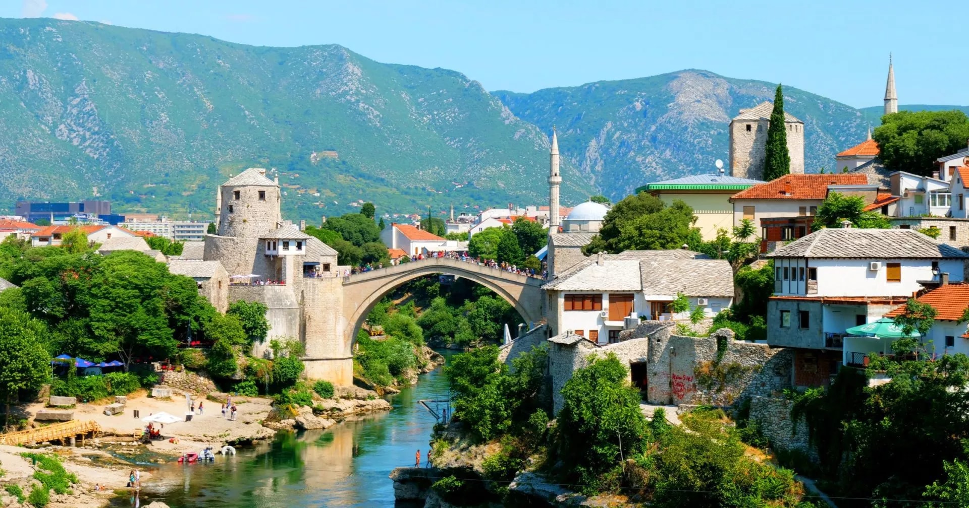 Mostar Stari Most bridge over turquoise river with mosque and green mountains in background.