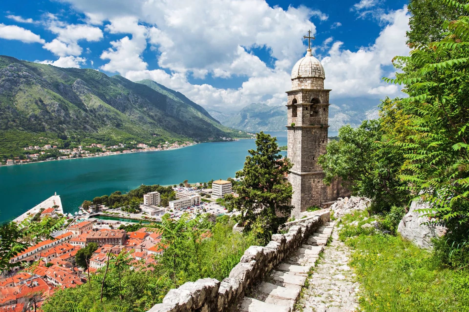 Stone bell tower overlooking Kotor Bay with steep green mountains and town below.