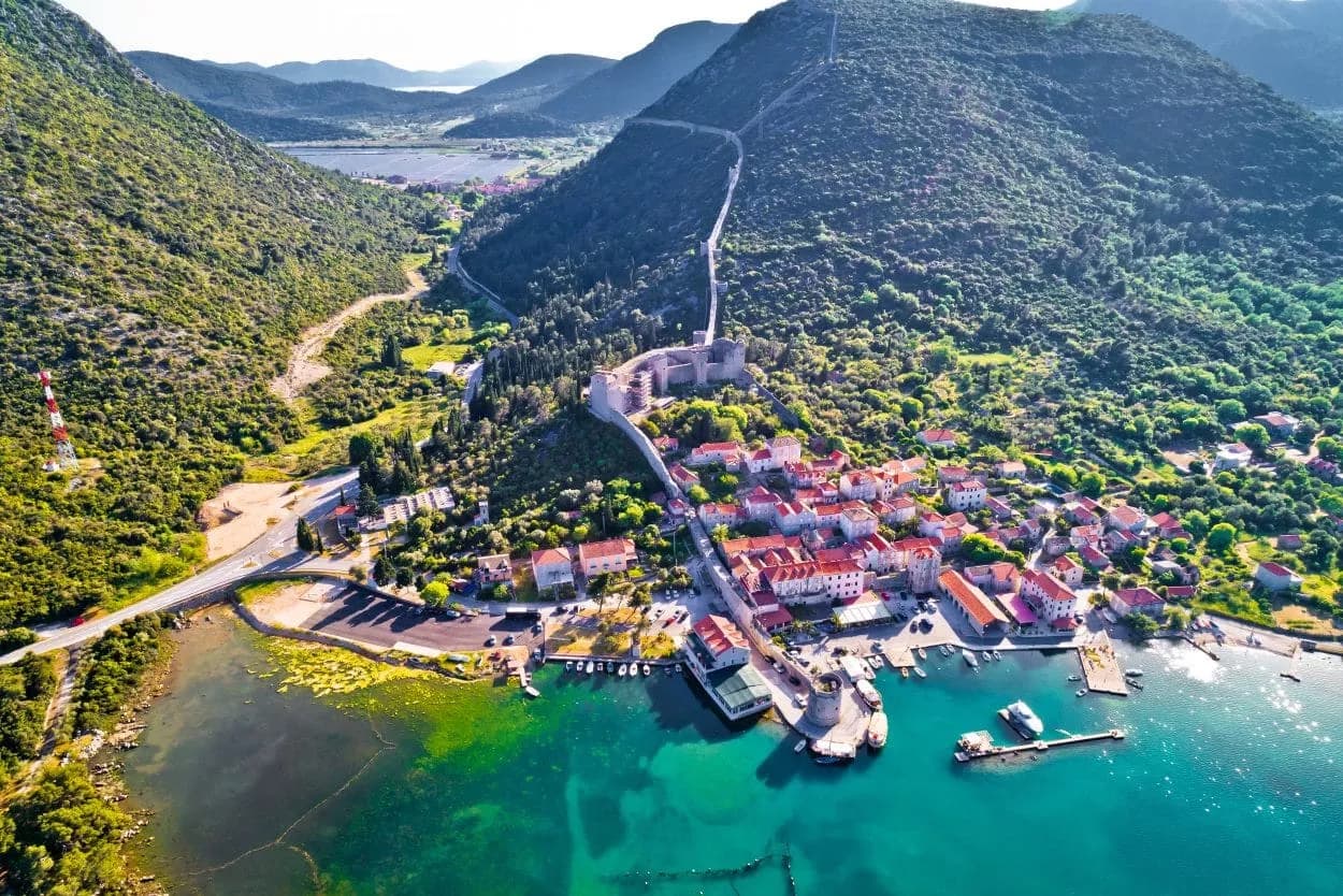 Coastal town with red-roofed buildings, stone walls, and turquoise water nestled between green mountains.
