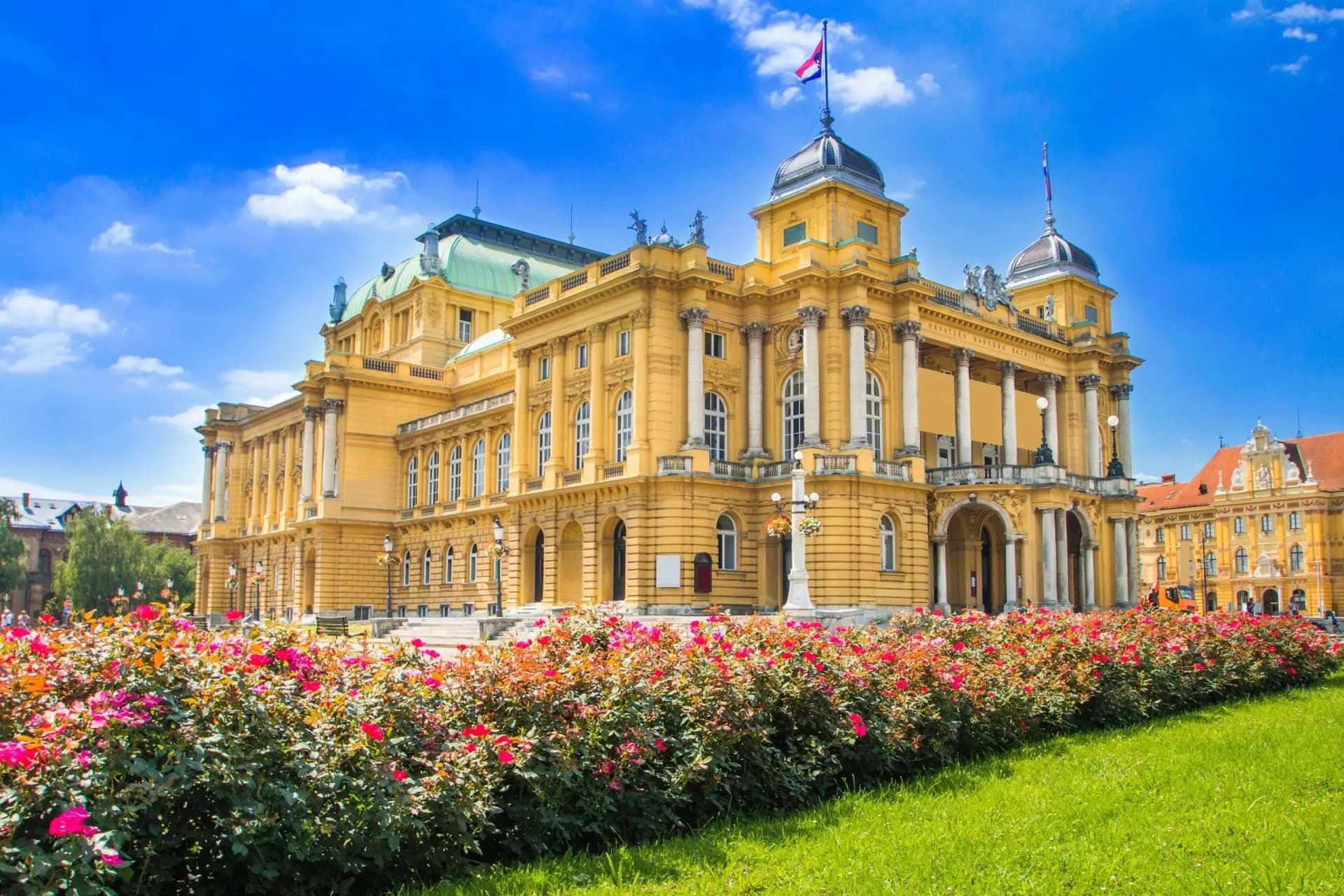 Croatian National Theater in Zagreb with vibrant rose bushes in the foreground under a blue sky.