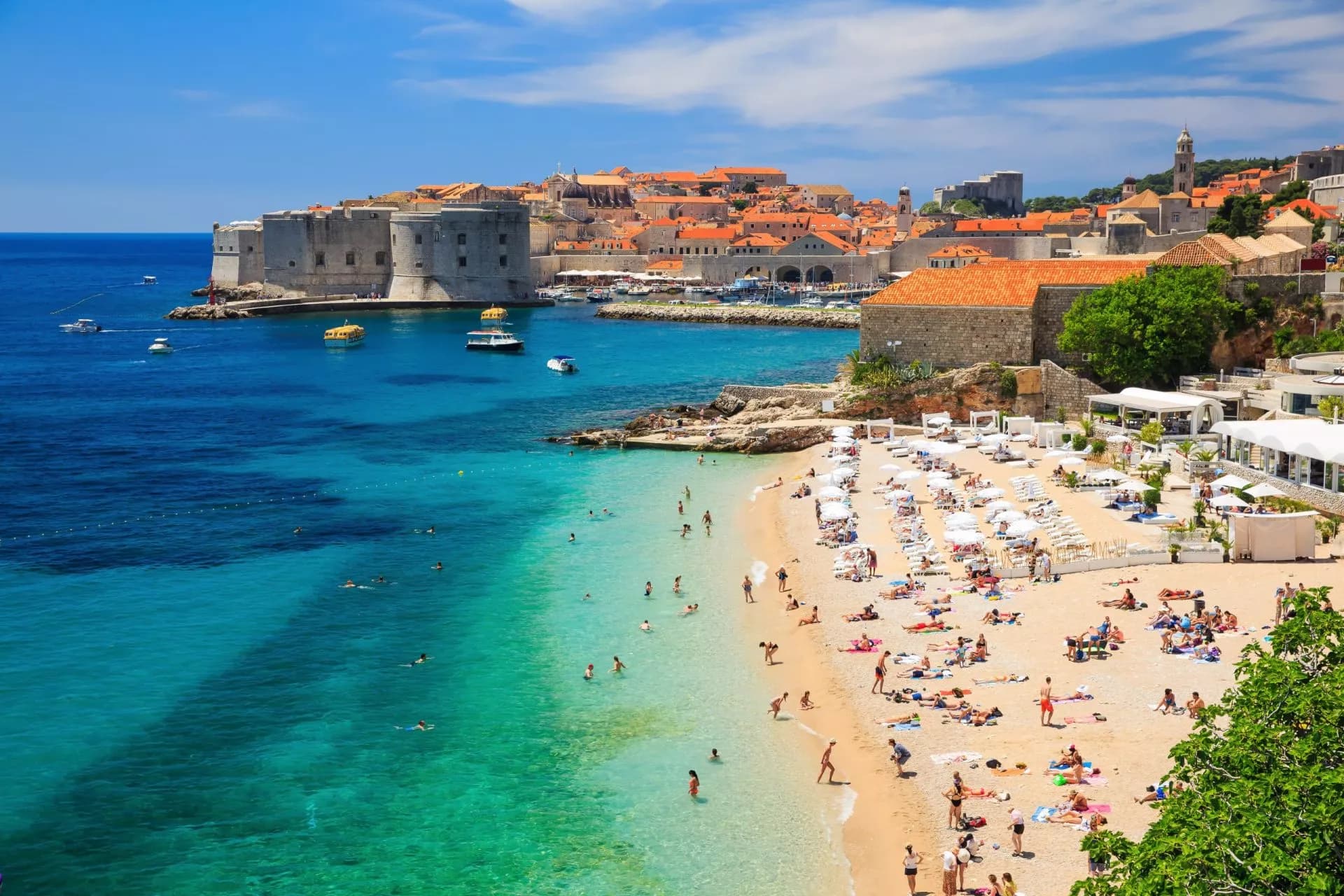 Beachgoers sunbathing near turquoise water with Dubrovnik's old town and fortress in the background.