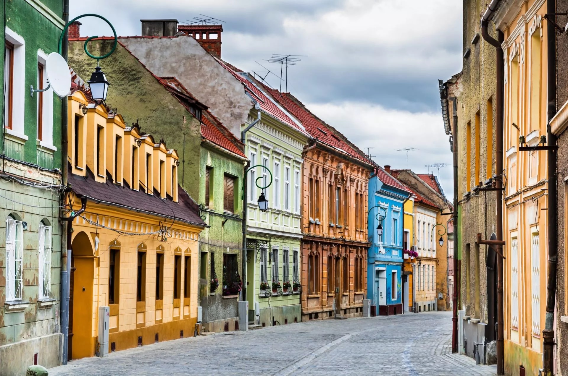 Cobblestone street lined with colorful historic buildings in Brasov, Romania, under a cloudy sky.