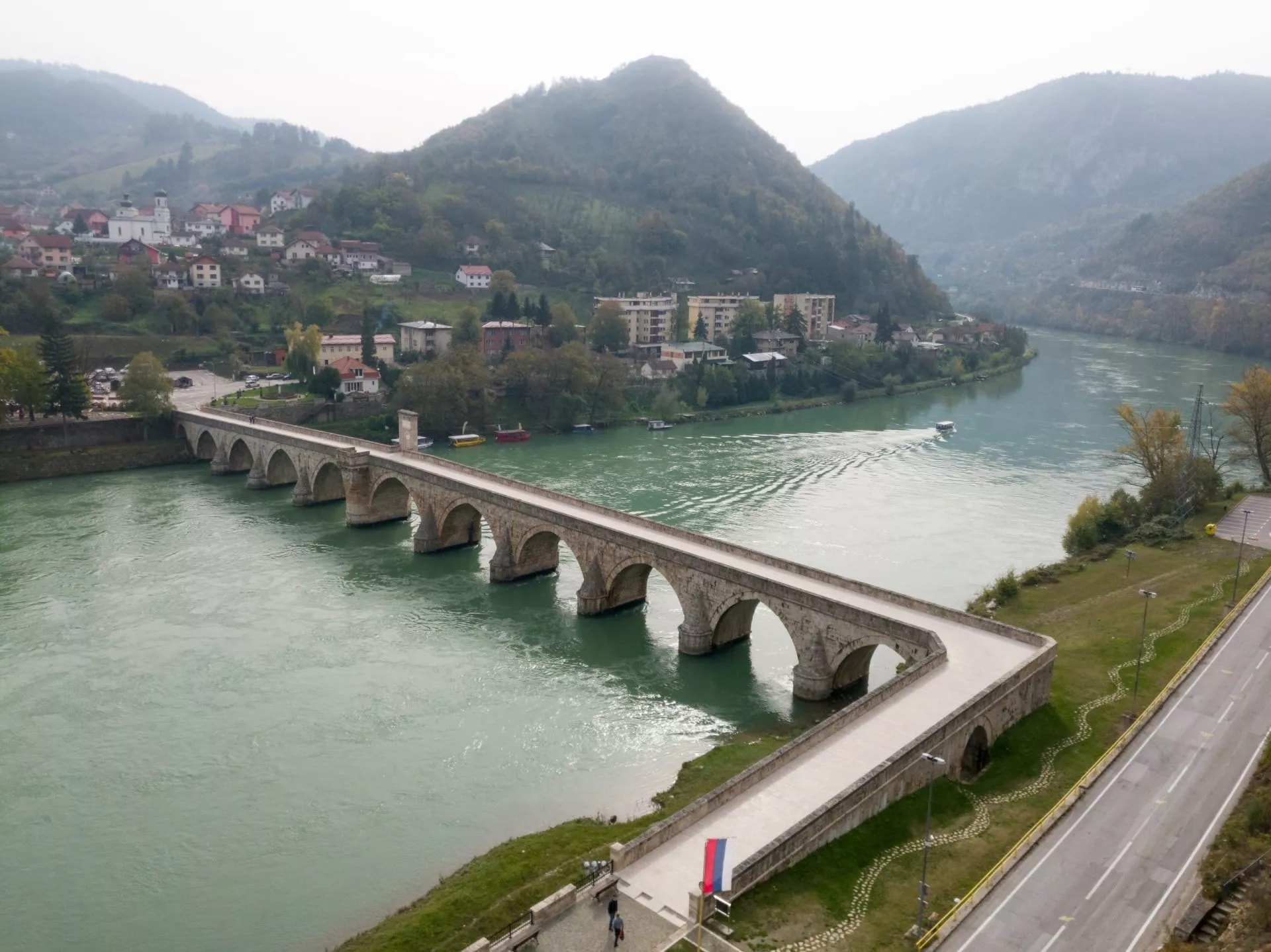 Old stone bridge over the green Drina River in Višegrad with a town on the hillside.