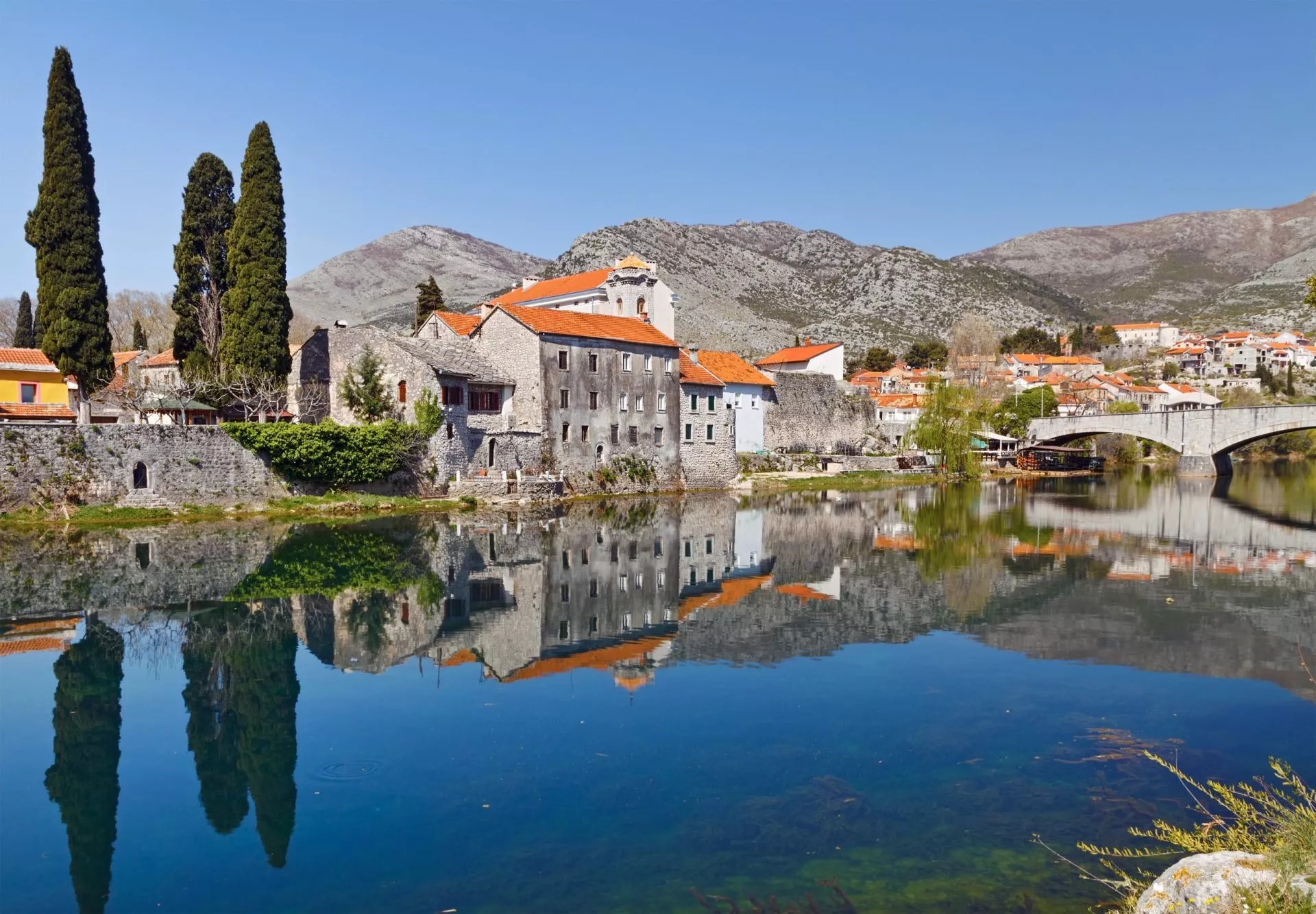Old town Trebinje stone buildings reflected in clear river water beneath mountains.