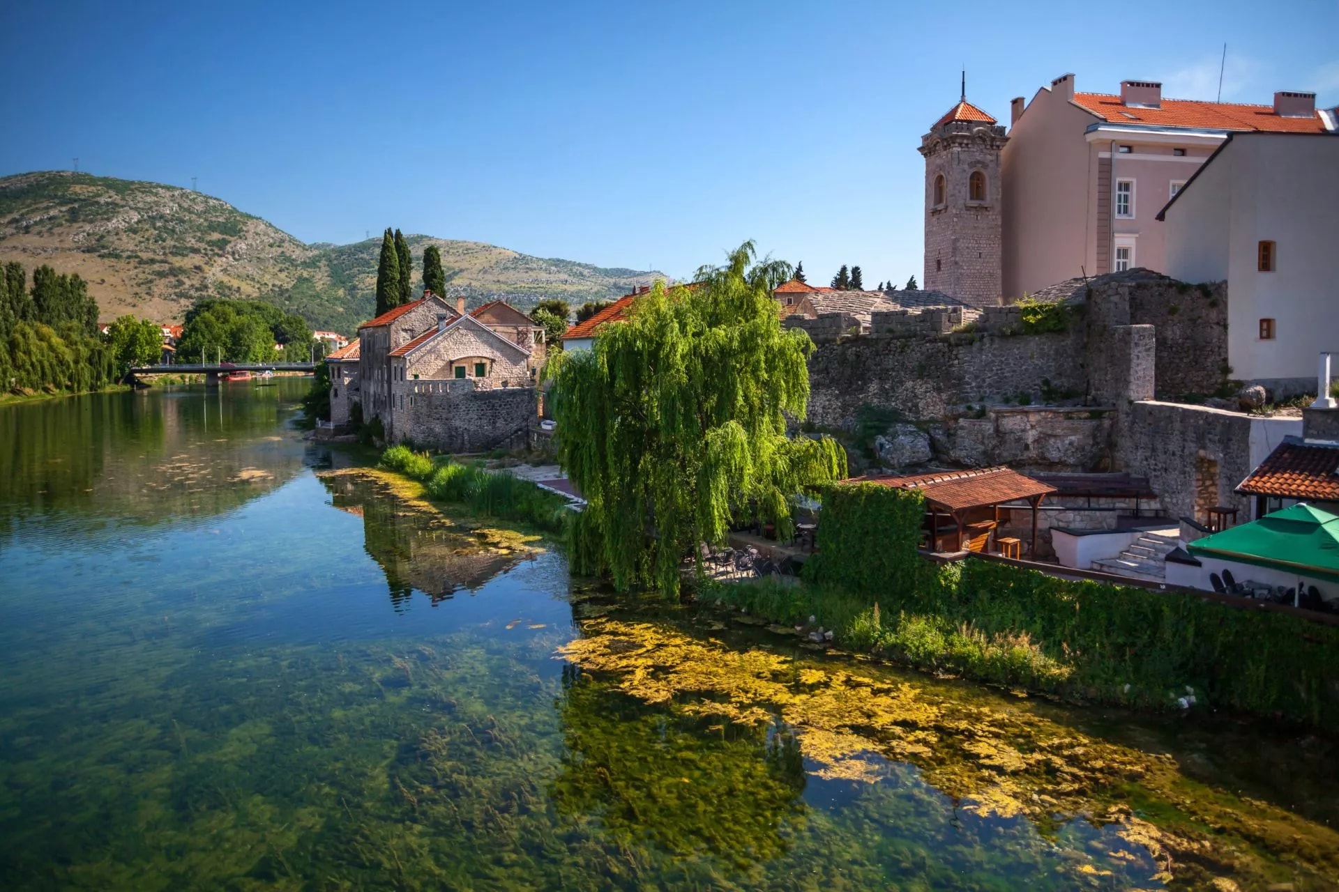 Old town Trebinje stone buildings along a clear river with green algae and mountains in the background.