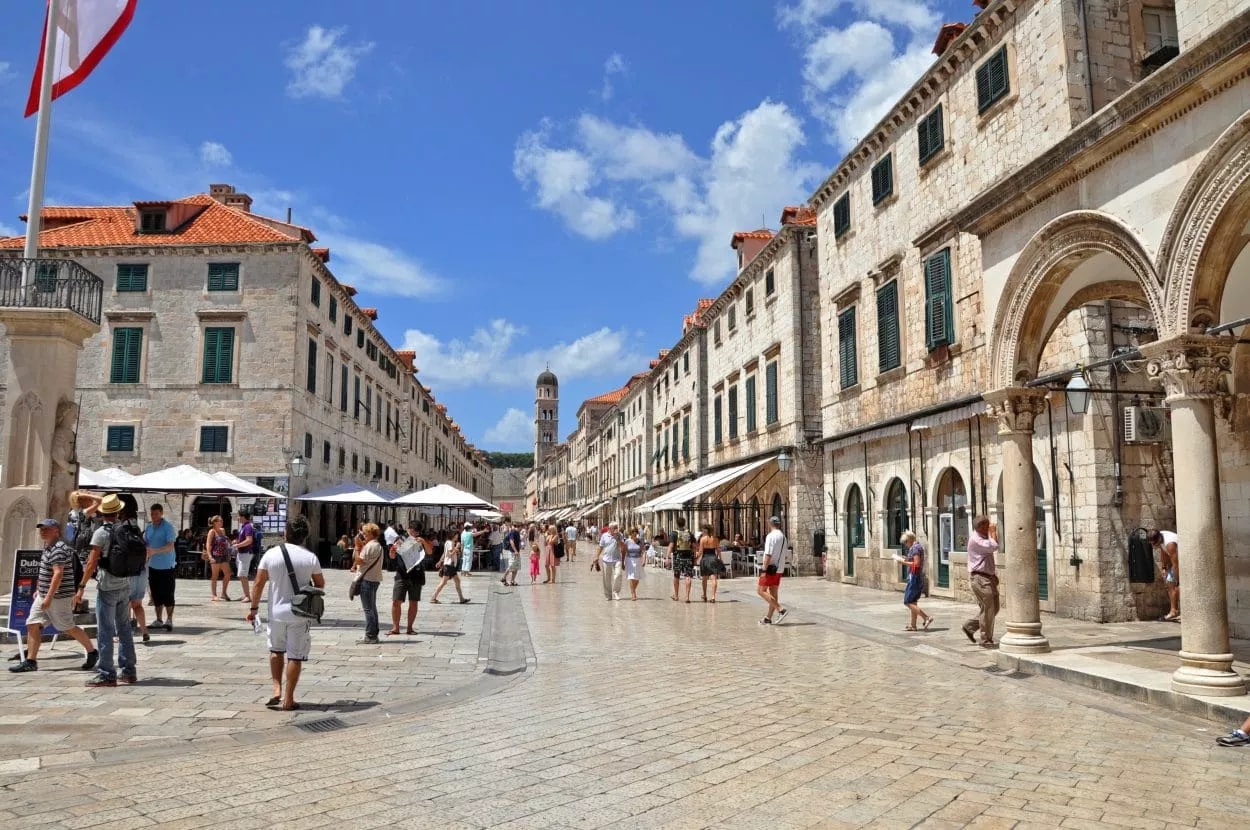 Pedestrians walking down the main stone street in Dubrovnik toward a distant bell tower.