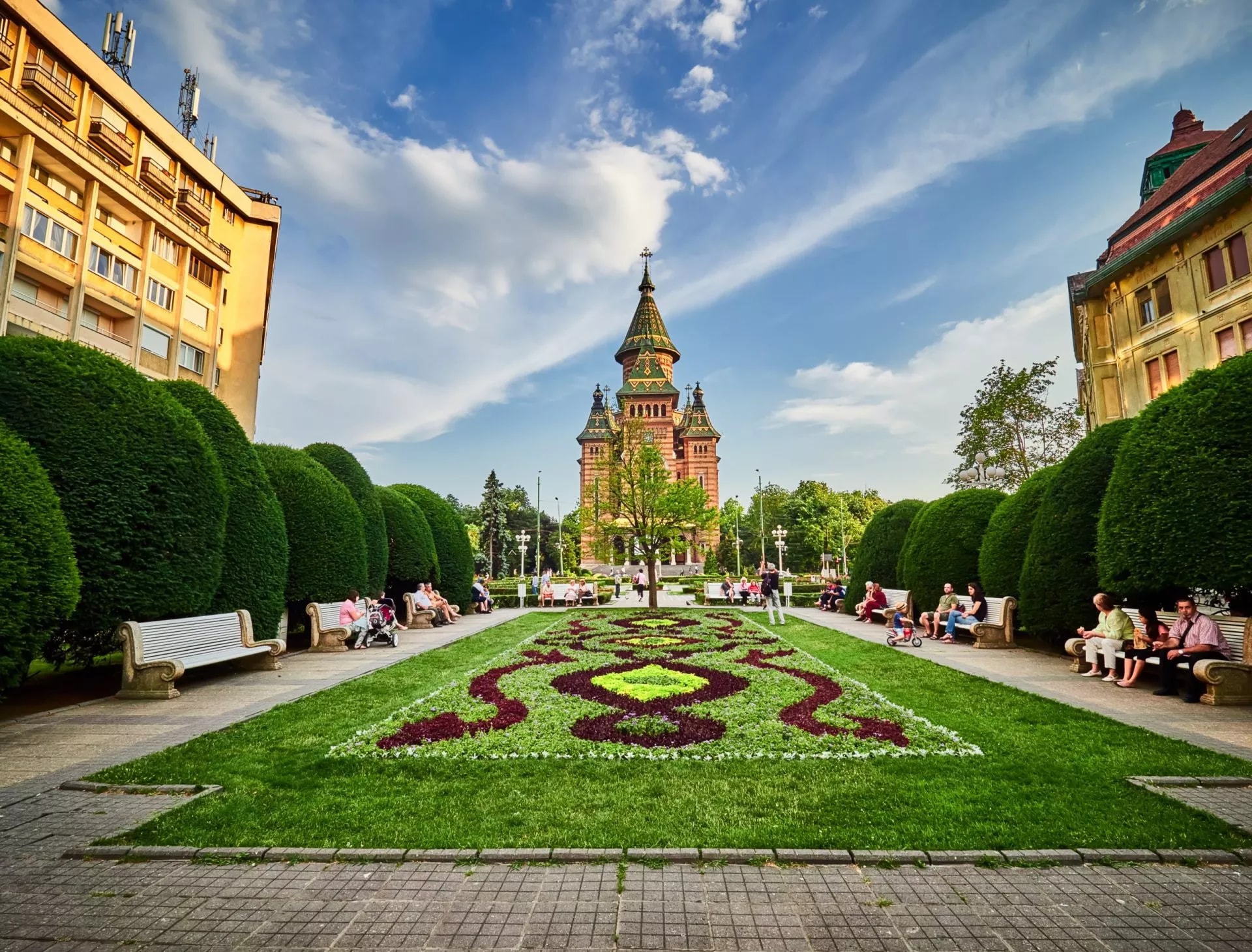 Timișoara Orthodox Cathedral viewed from a park with flowerbeds and manicured hedges.