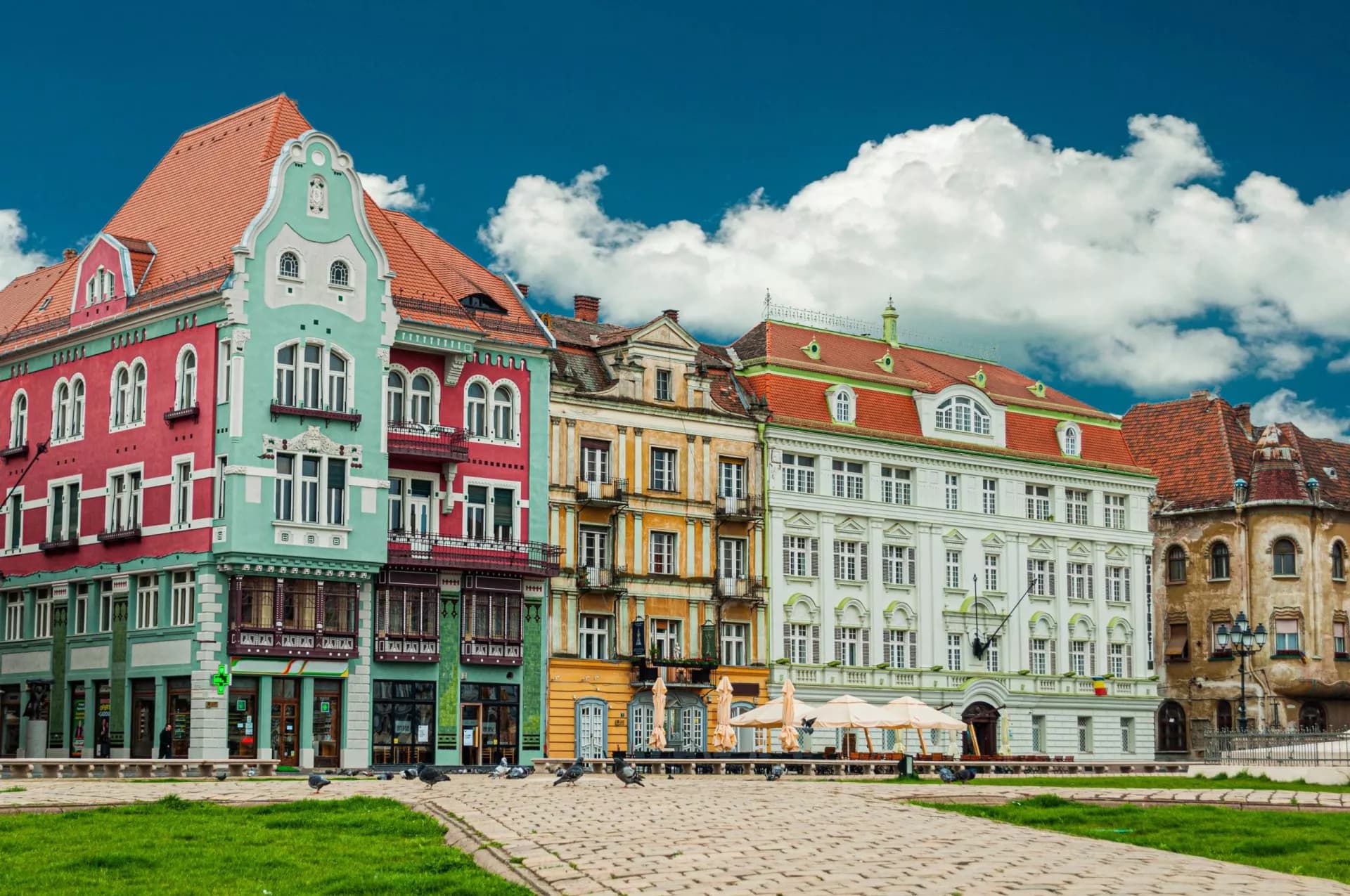 Colorful historic buildings line Unirii Square in Timisoara under a bright blue, cloudy sky.