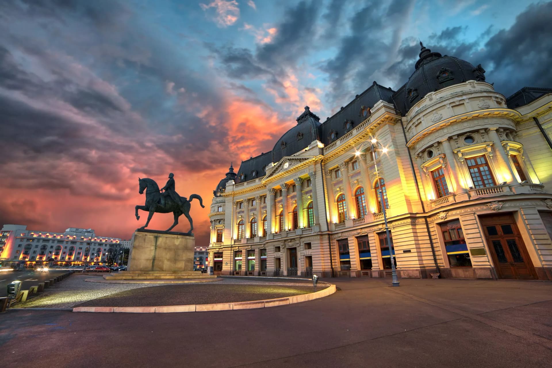 Equestrian statue outside illuminated University Library by night in Bucharest under dramatic sunset sky.