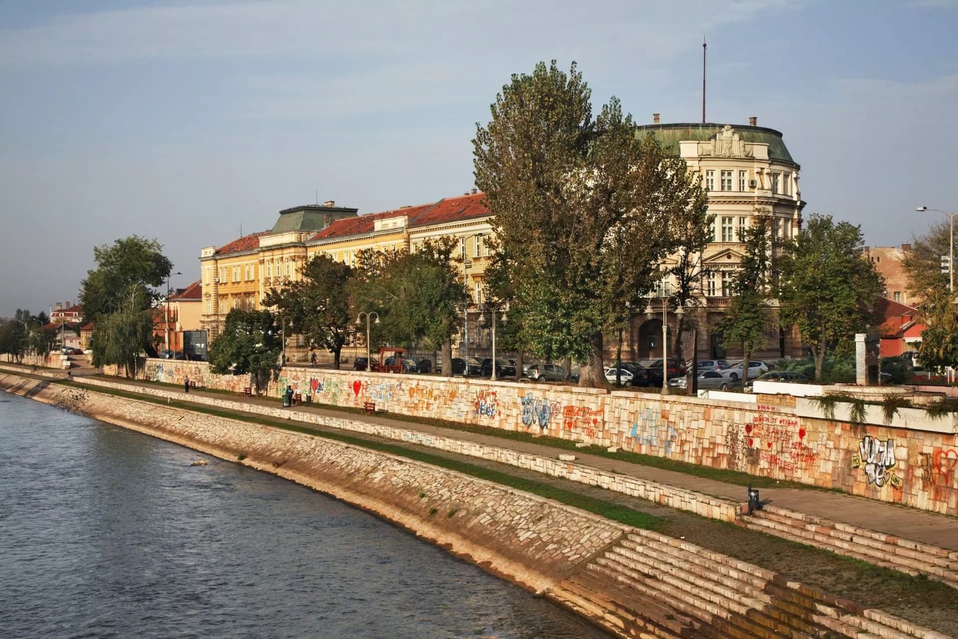 Embankment of Nišava River in Niš, Serbia, with historic buildings and graffiti wall.
