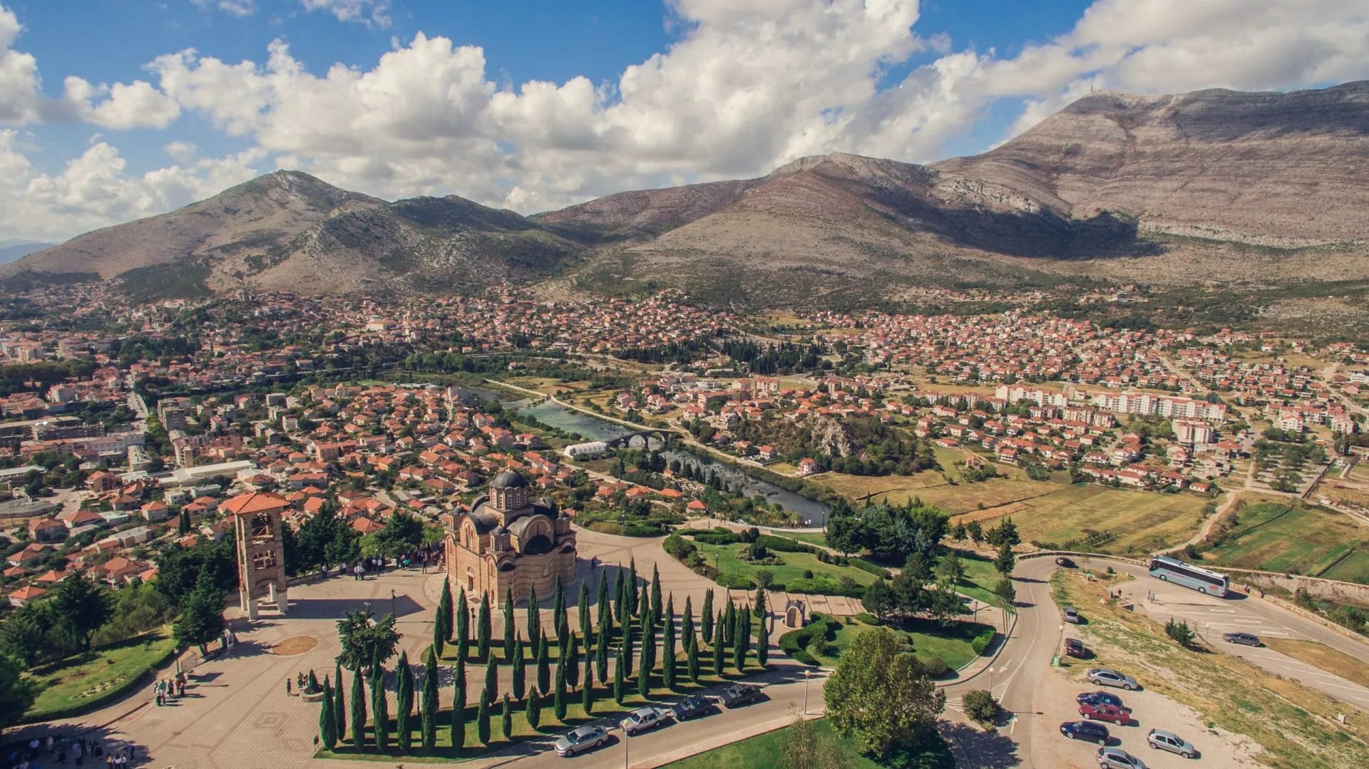 Aerial view of Trebinje city with Orthodox church, river, and surrounding mountains under cloudy sky.