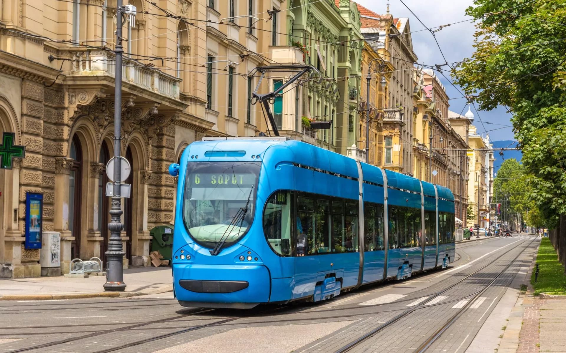 Modern blue tram on street tracks in Zagreb, Croatia, with historic buildings and trees.