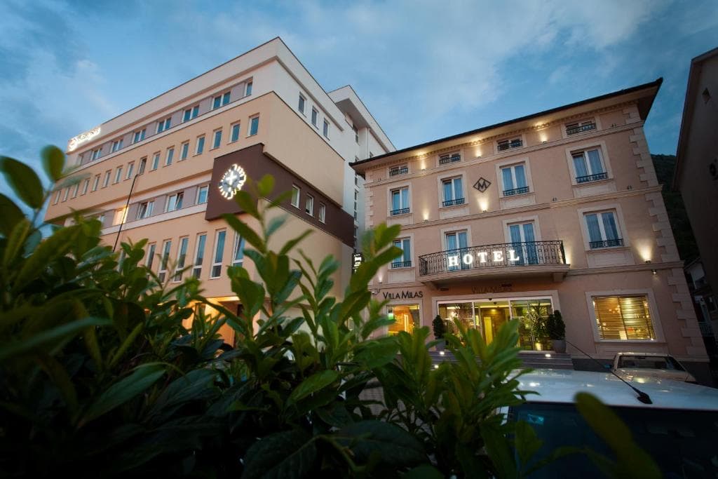 Hotel Villa Milas exterior with illuminated signage seen through dark green foliage at dusk.