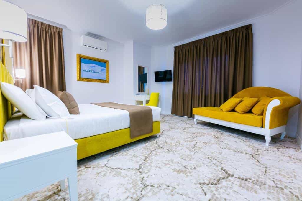 Hotel bedroom with yellow bed, white linens, yellow chaise lounge, and brown curtains.