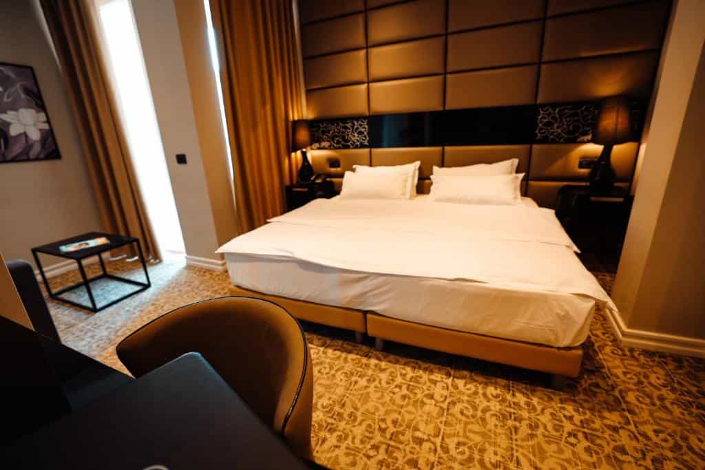Hotel Senator Dve bedroom with large white bed, padded headboard, and window with gold curtains.