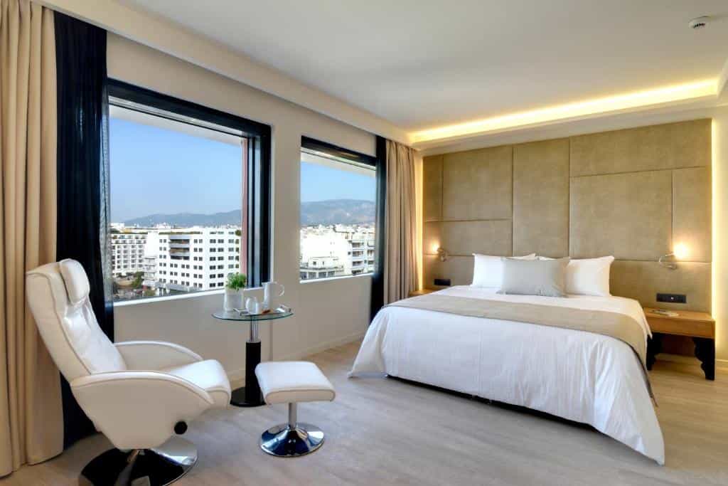 Hotel room with white bedding, modern chair, and city view from window, Athens.