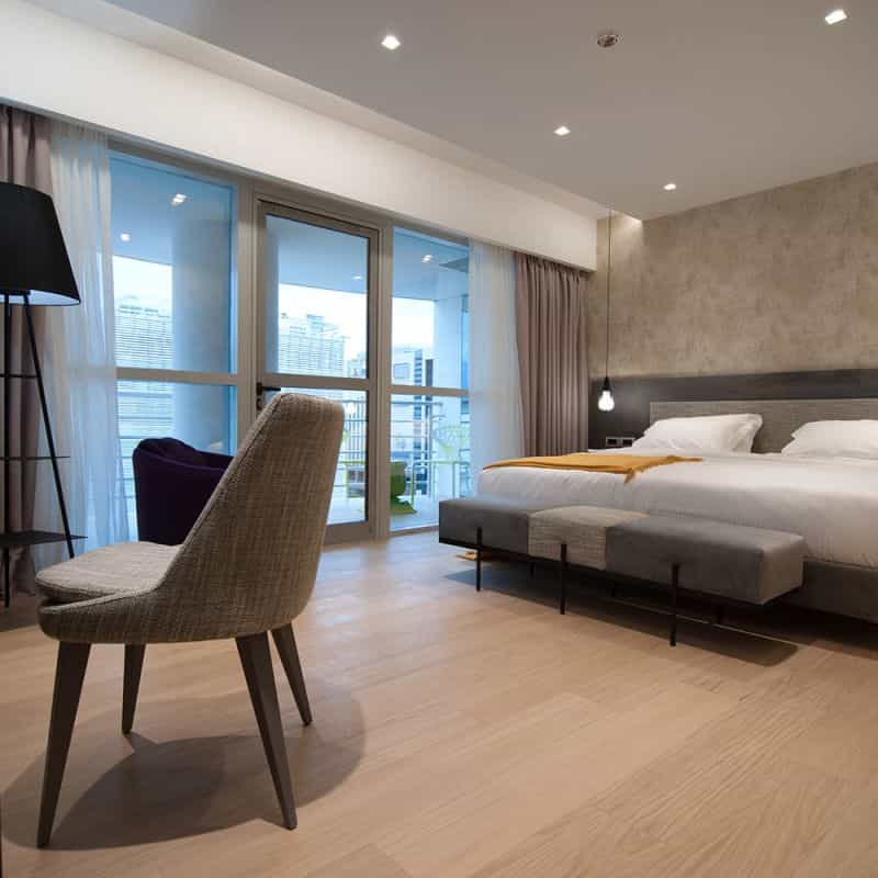 Modern hotel bedroom with large window view of city buildings and light wood floor.